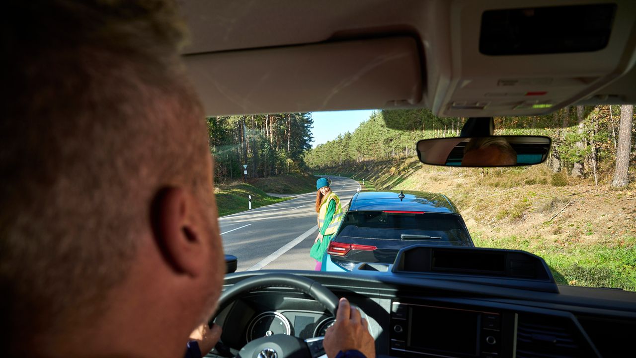 Ein Mann parkt seinen Volkswagen hinter einem anderen Volkswagen. Durch die Windschutzscheibe sieht man eine Frau in Warnweste, die am anderen Fahrzeug angelehnt ist.