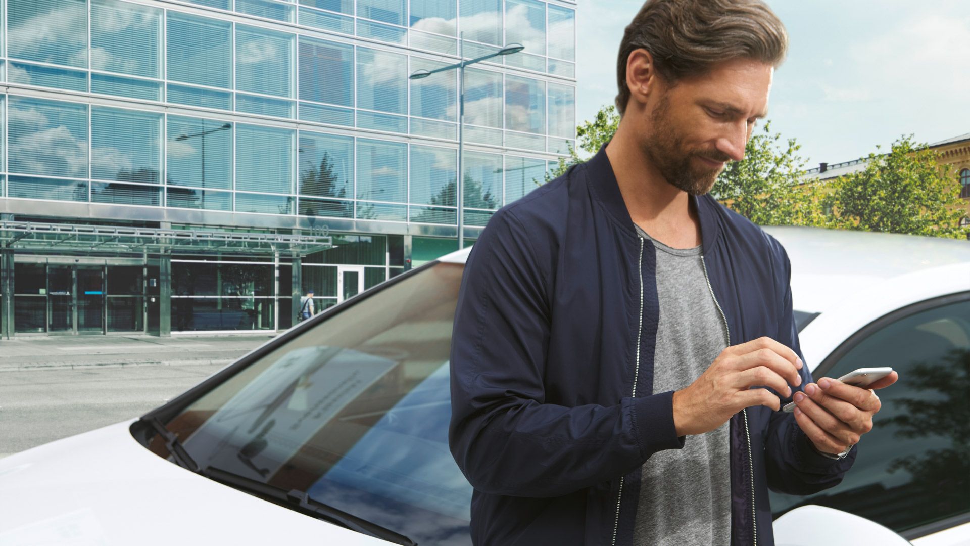 Ein Mann tippt in sein Smartphone. Im Hintergrund ein weißer Volkswagen vor einem Gebäude.