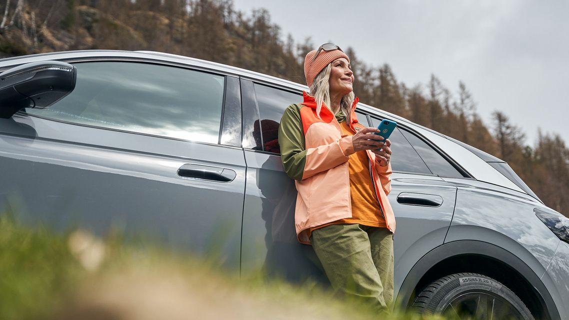 Eine Frau  mit Smartphone lehnt an einem Volkswagen und blickt in die Ferne