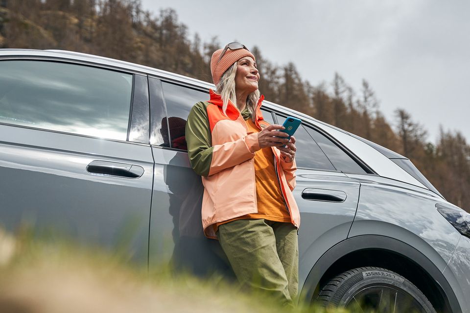 Eine Frau  mit Smartphone lehnt an einem Volkswagen und blickt in die Ferne