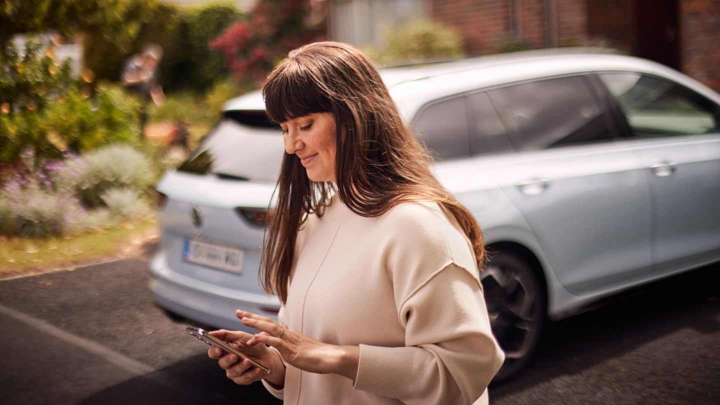 Frau steht vor VW Golf Variant und nutzt die Volkswagen App auf ihrem Smartphone