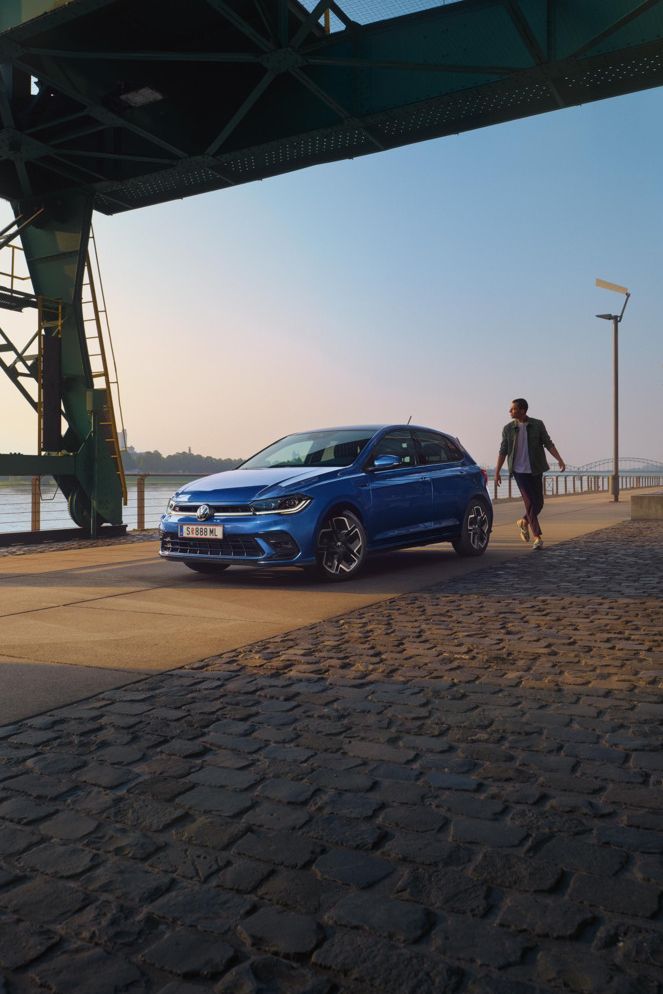  Ein blauer VW Polo R-Line steht an der Promenade neben einem Fluss, ein Mann läuft um das Auto herum.