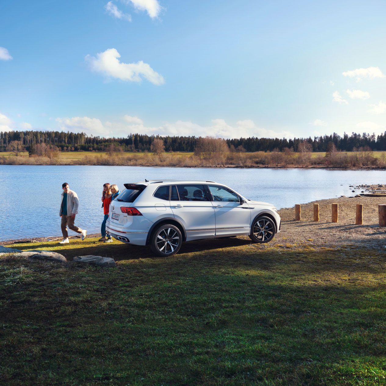 Ein weißer VW Tiguan Allspace steht geparkt auf einer Wiese vor einem See. Dahinter laufen 3 Menschen entlang.