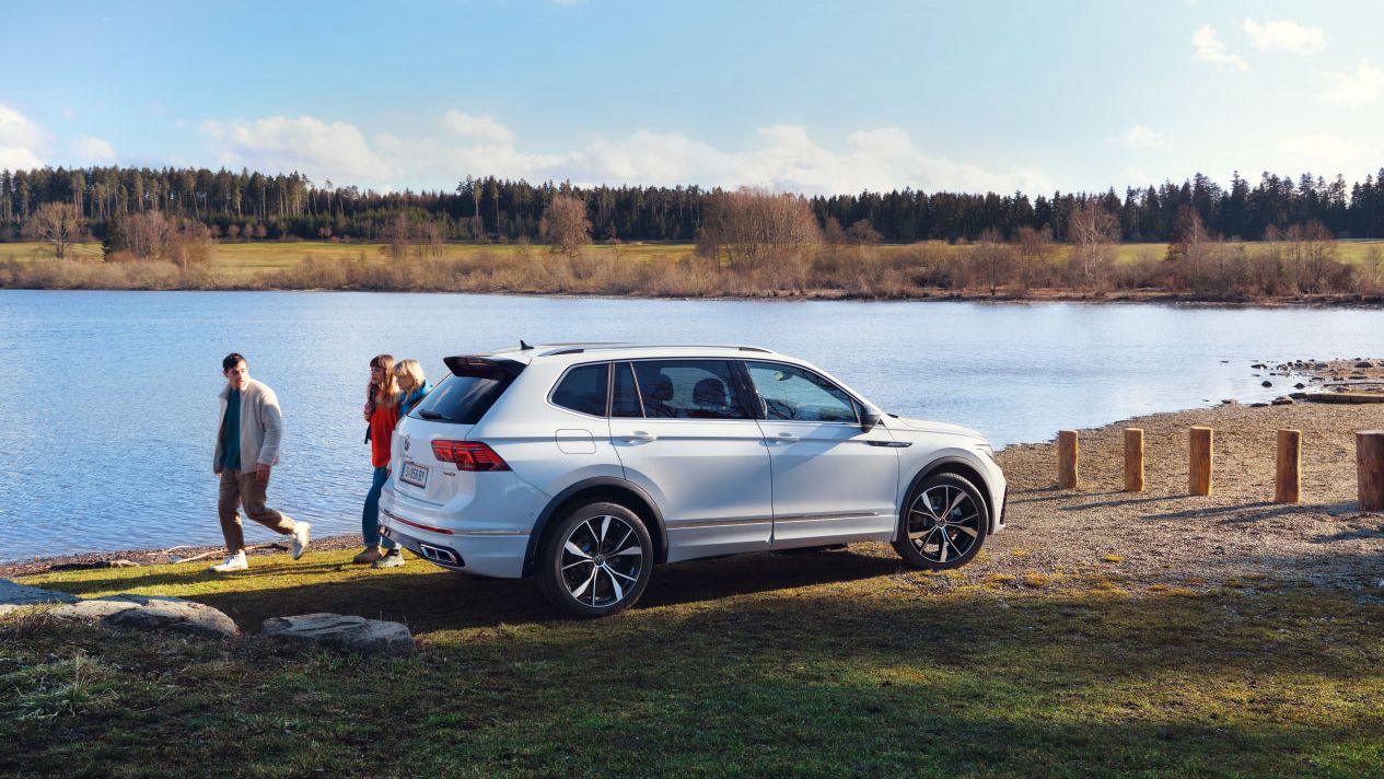 Ein weißer VW Tiguan Allspace steht geparkt auf einer Wiese vor einem See. Dahinter laufen 3 Menschen entlang.