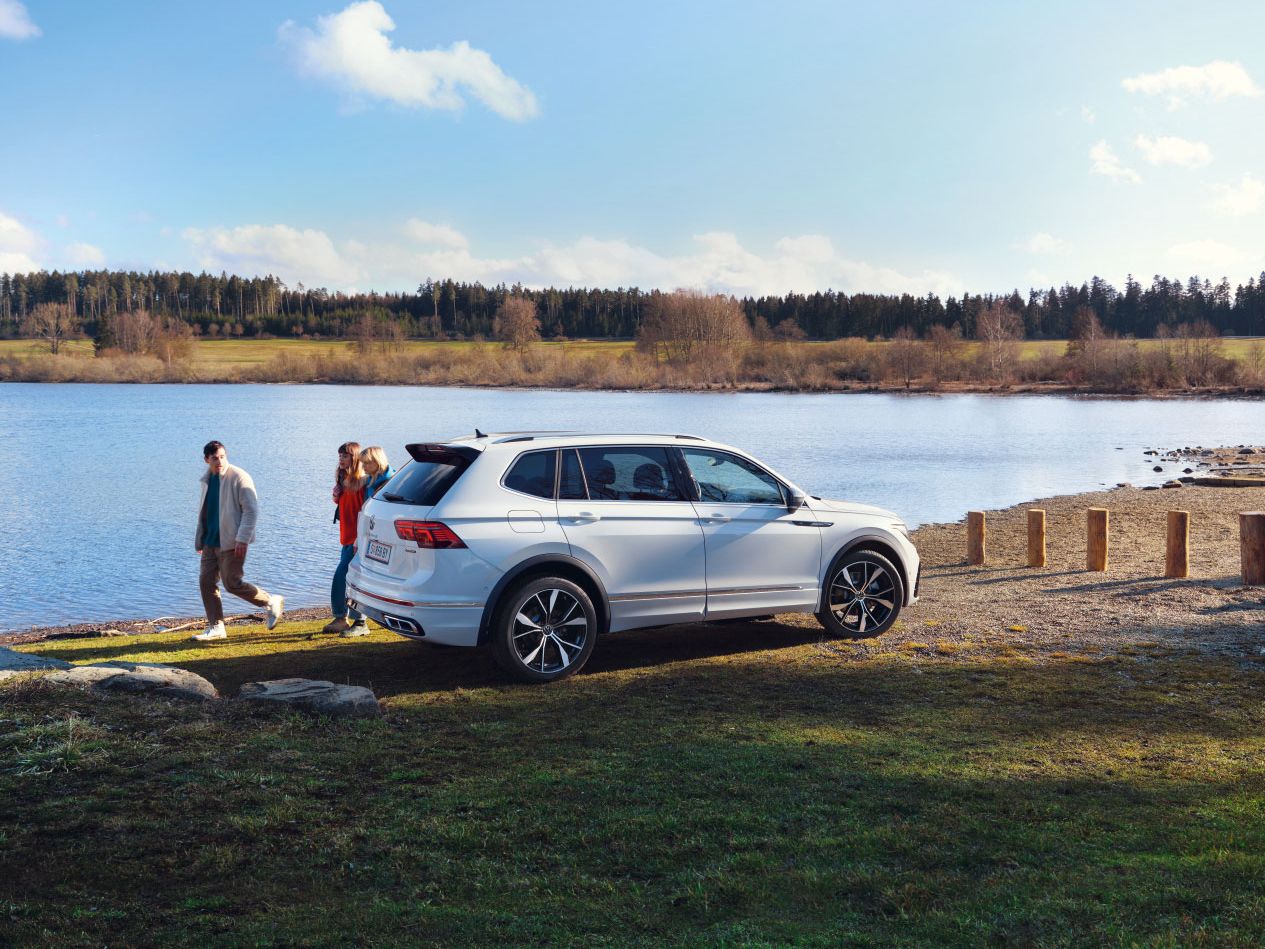 Ein weißer VW Tiguan Allspace steht geparkt auf einer Wiese vor einem See. Dahinter laufen 3 Menschen entlang.