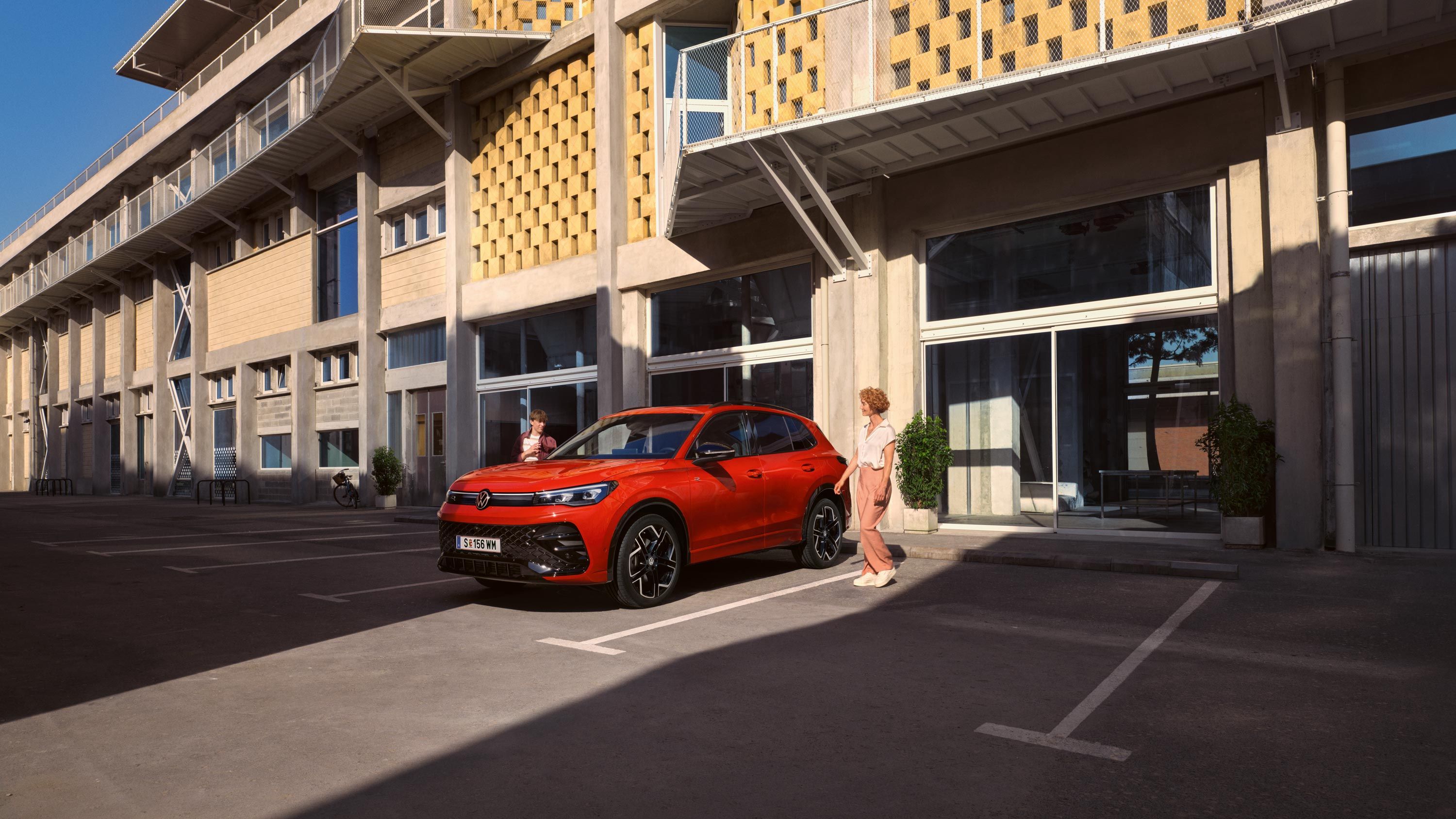 Roter VW Tiguan R-Line in Seitenansicht steht vor einem Gebäude, eine junge Frau geht auf der Fahrerseite vorbei.