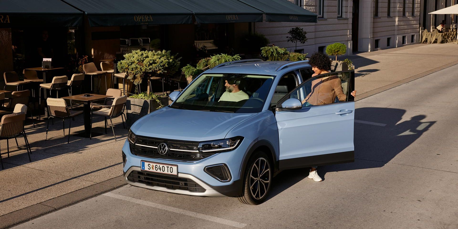 Eine Frau steigt auf der Fahrerseite in den blauen VW T-Cross Style, der vor einem urbanen Straßencafé parkt.