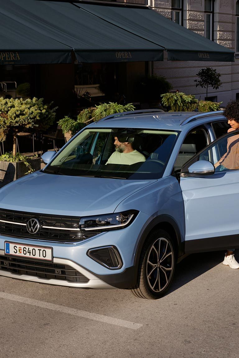 Eine Frau steigt auf der Fahrerseite in den blauen VW T-Cross Style, der vor einem urbanen Straßencafé parkt.