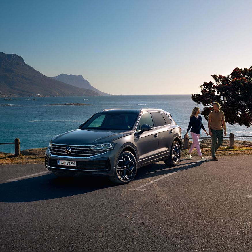 Der neue Volkswagen Touareg auf einem Parkplatz an der Küste, an ihm gehen eine Frau und ein Mann vorbei