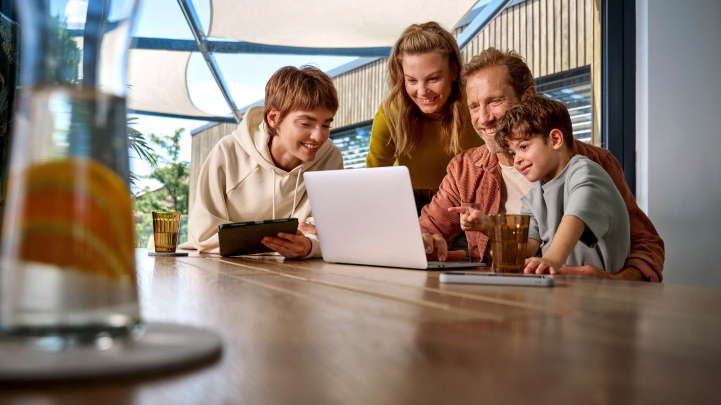 Eine Familie ist um einen Tisch versammelt und schaut fröhlich auf einen Laptop-Bildschirm