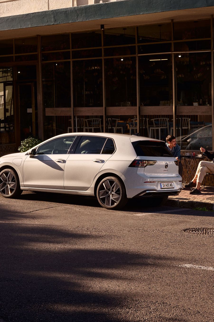 Zwei Männer sitzen vor einem Cafe und unterhalten sich. Vor ihnen ein neuer Golf 8.