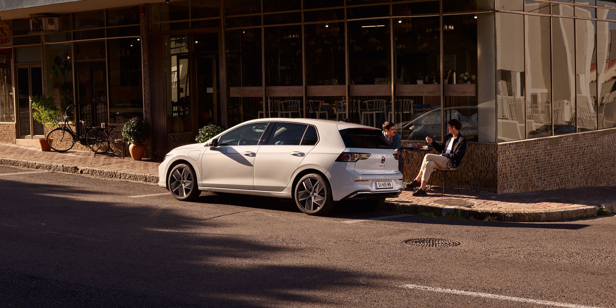 Zwei Männer sitzen vor einem Cafe und unterhalten sich. Vor ihnen ein neuer Golf 8.