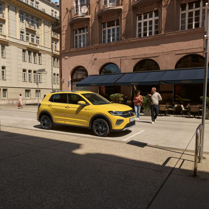 Ein gelber VW T-Cross ist in einer Stadt geparkt, hinter ihm sieht man Menschen