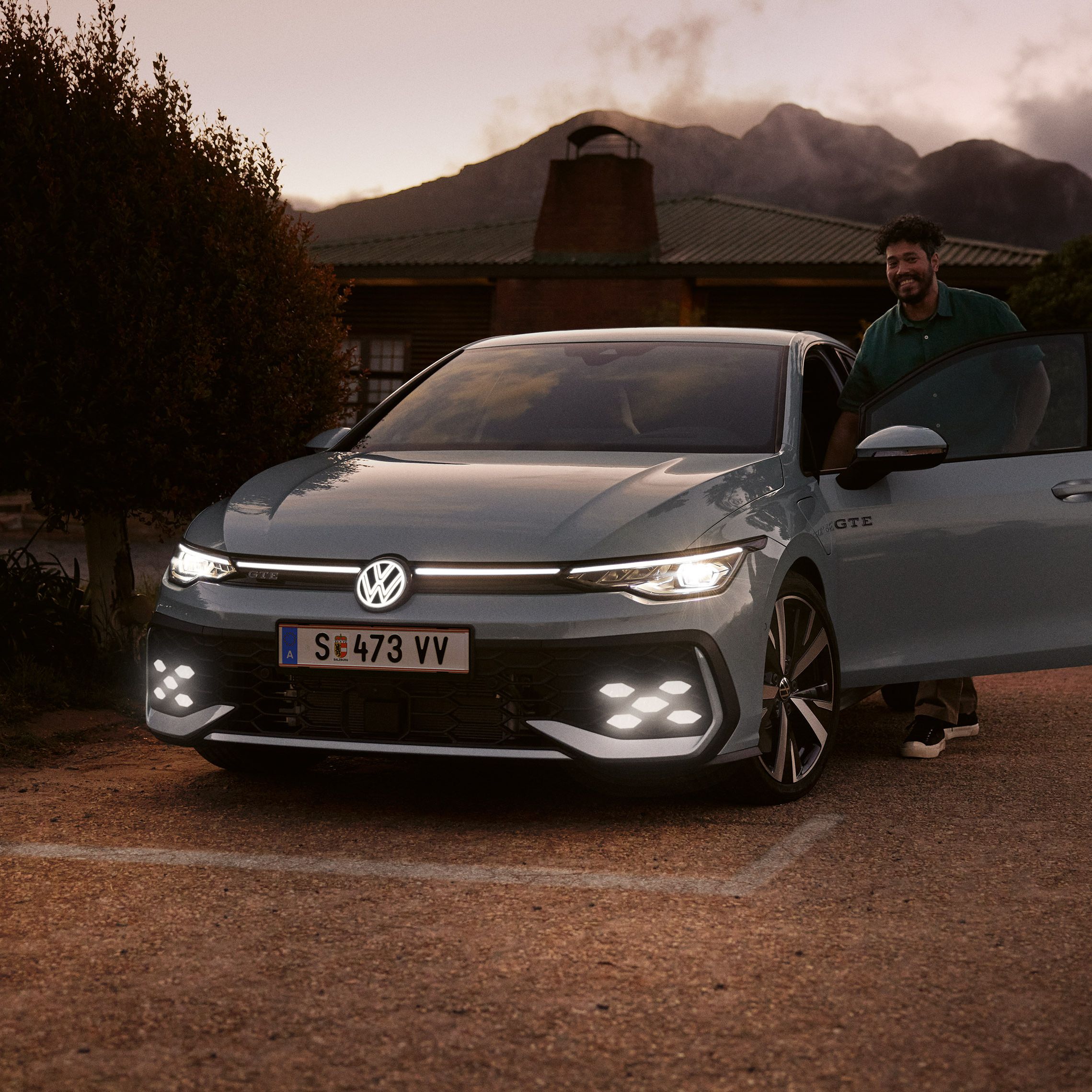 Frontansicht eines grauen VW Golf GTE mit IQ. Light LED Matrix Scheinwerfern in abendlicher Stimmung, Mann steigt in Golf GTE