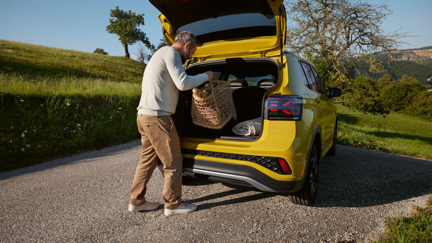 Ein Mann stellt einen großen Picknickkorb in den Kofferraum eines gelben VW T-Cross, der auf einer Straße in hügeliger Landschaft steht.