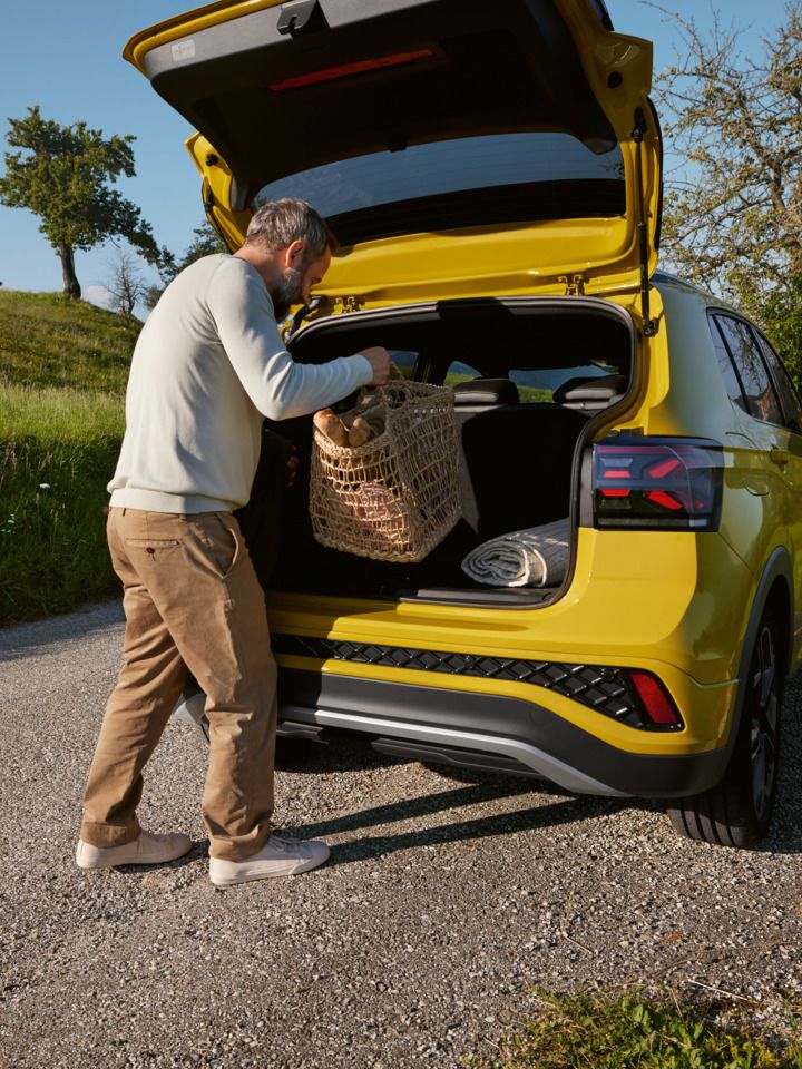 Ein Mann stellt einen großen Picknickkorb in den Kofferraum eines gelben VW T-Cross, der auf einer Straße in hügeliger Landschaft steht.