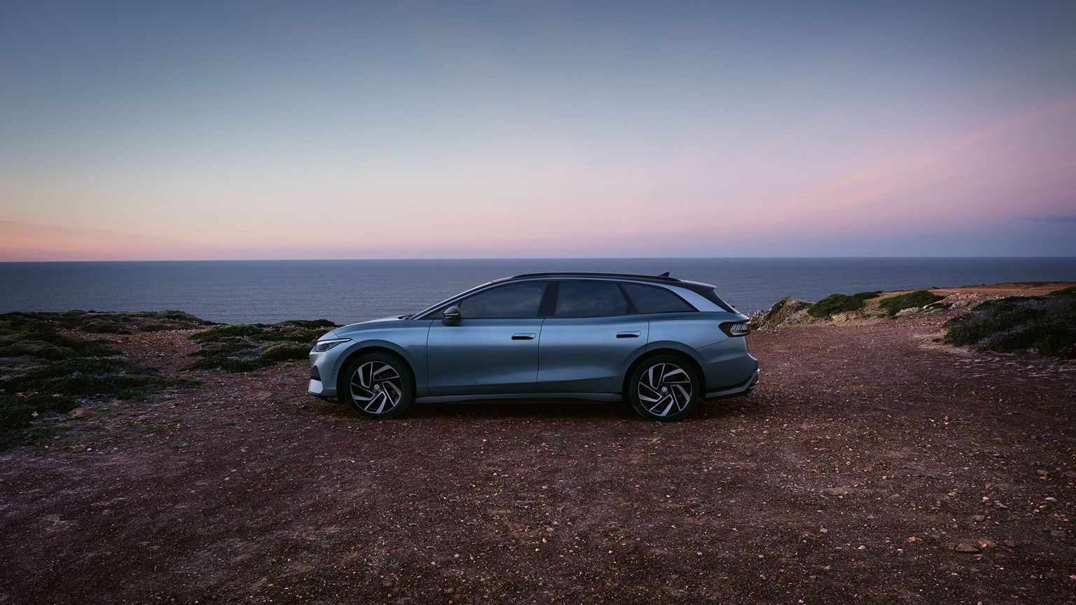 Seitenansicht des VW ID.7 in der Farbe Stonewashed Blue geparkt an der Küste mit Sonnenuntergang