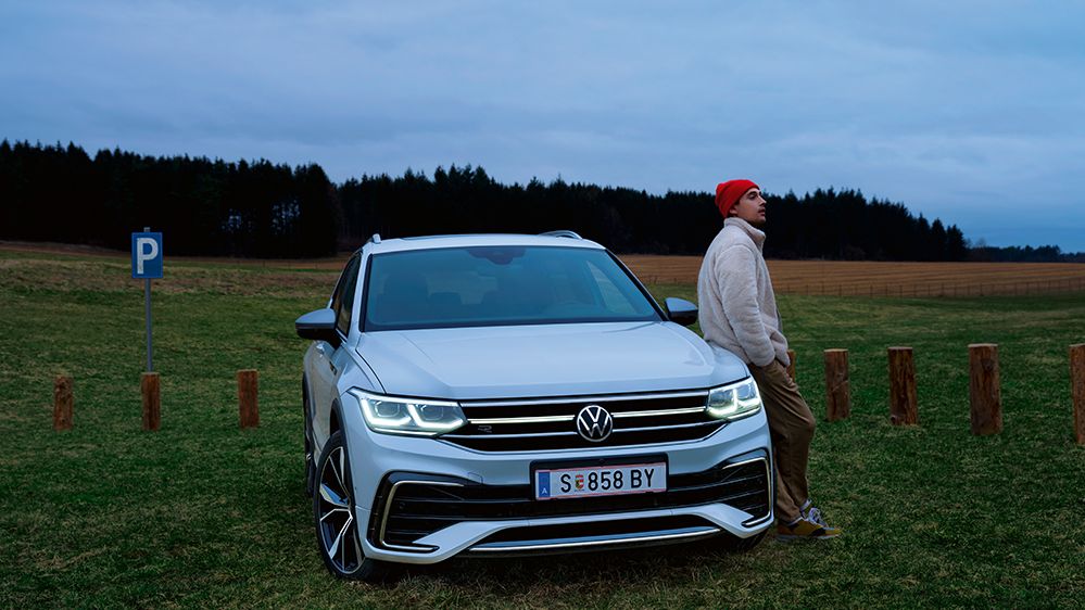 Ein weißer Tiguan Allspace ist geparkt auf einer Wiese, ein Mann lehnt sich an die Front des Autos an