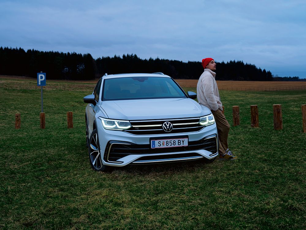 Ein weißer Tiguan Allspace ist geparkt auf einer Wiese, ein Mann lehnt sich an die Front des Autos an