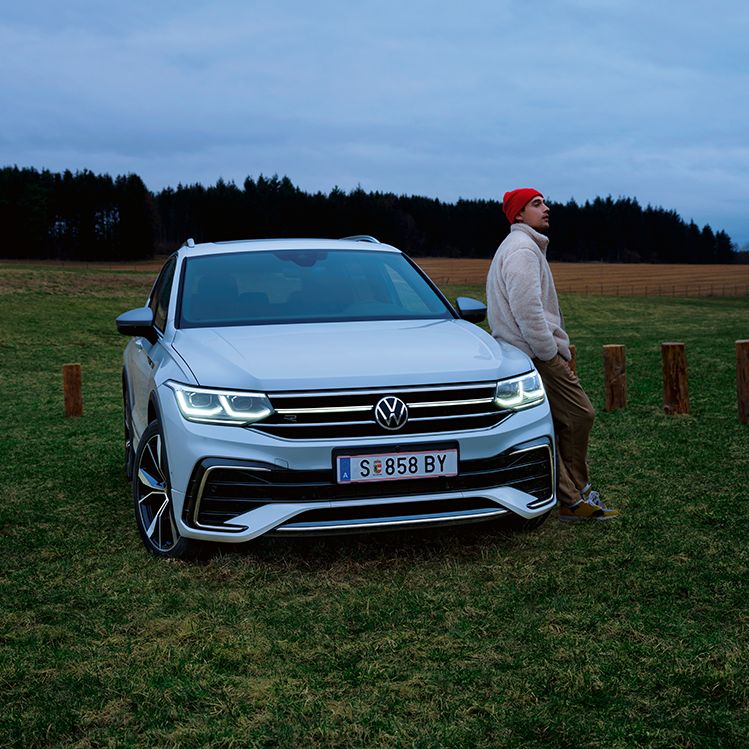 Ein weißer Tiguan Allspace ist geparkt auf einer Wiese, ein Mann lehnt sich an die Front des Autos an