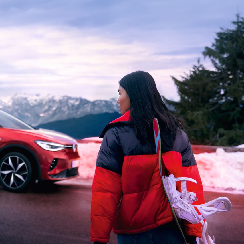 Seitenansicht eines roten VW ID.4 GTX vor Bergkulisse, im Vordergrund blickt eine Frau zum Auto