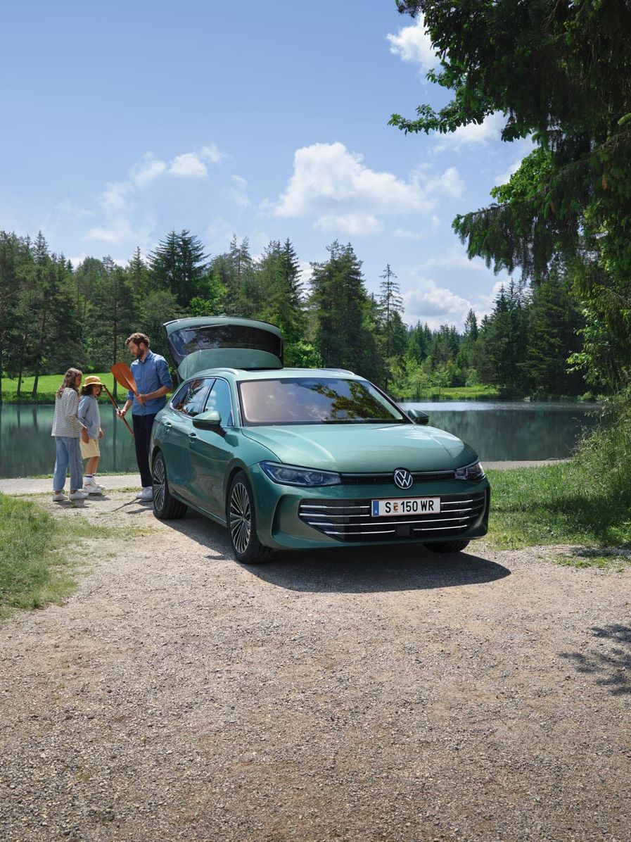 Der neue VW Passat Elegance in Maripositgrün Metallic parkt an einem Badesee mit geöffnetem Kofferraum. Ein Mann verteilt Freitzeitspielzeug an zwei Kinder.