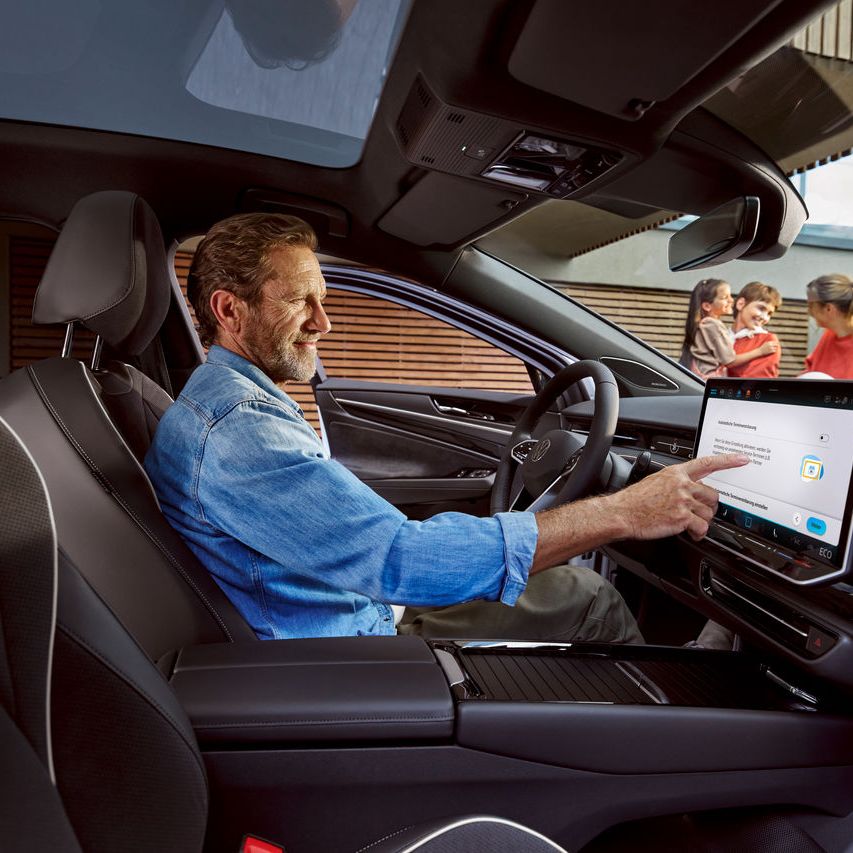 Mann sitzt in seinem Volkswagen und berührt das Display, im Hintergrund seine Familie