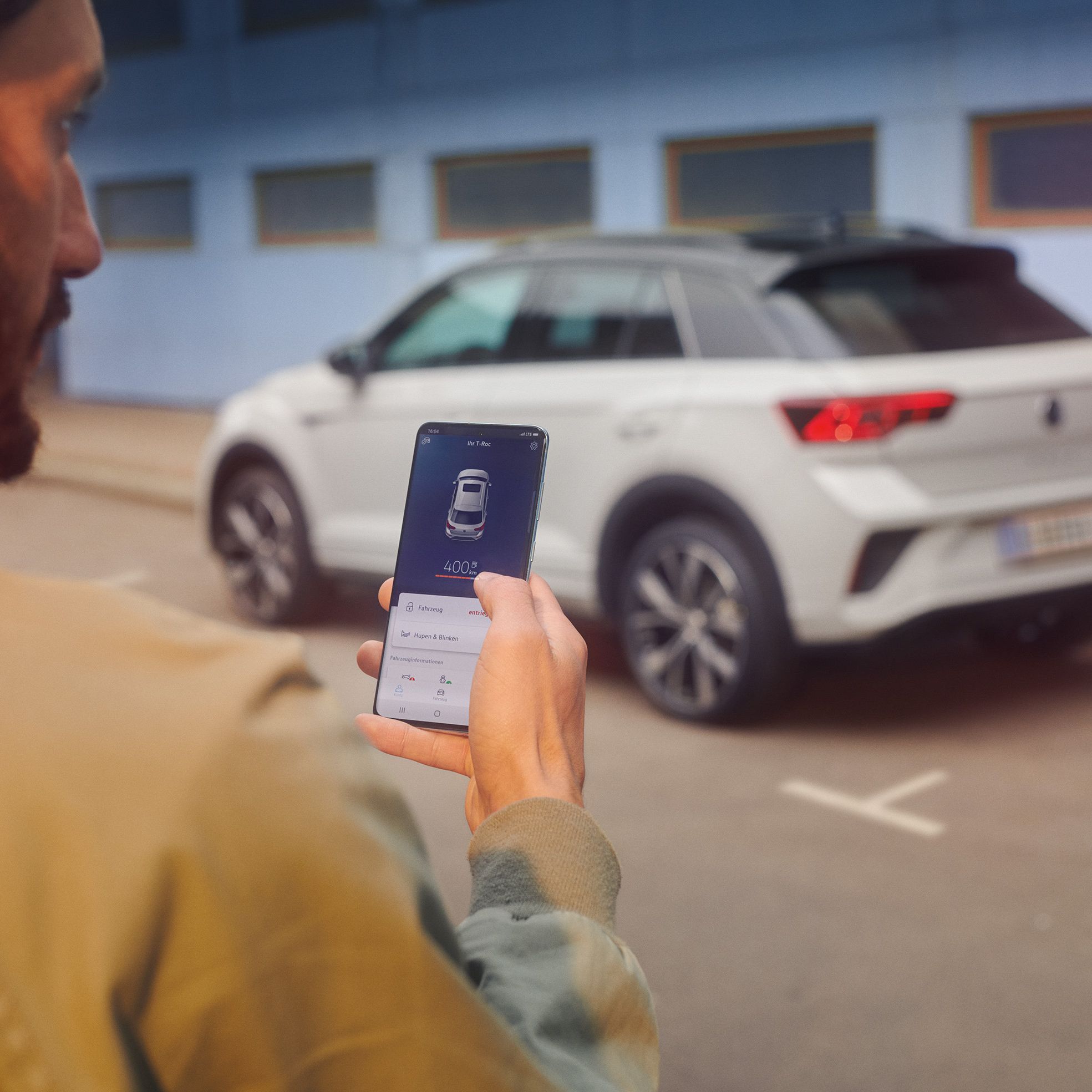 VW T-Roc R-Line in weiß vor einem Gebäude, Seite sichtbar, Mann steht mit Smartphone im Vordergrund