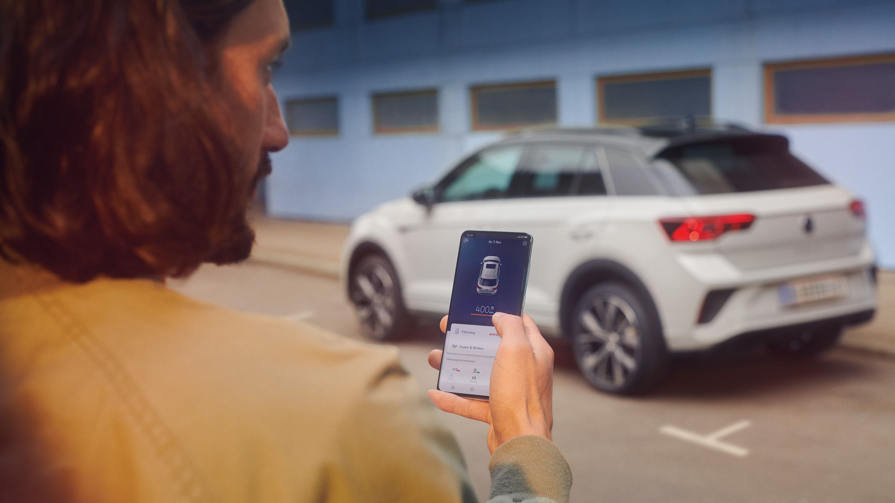 VW T-Roc R-Line in weiß vor einem Gebäude, Seite sichtbar, Mann steht mit Smartphone im Vordergrund