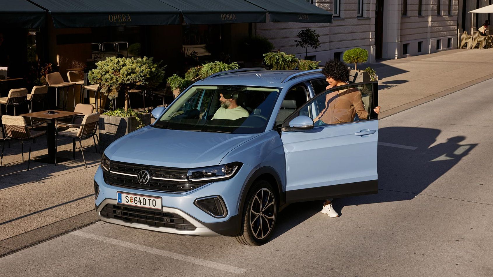 Eine Frau steigt auf der Fahrerseite in den blauen VW T-Cross Style, der vor einem urbanen Straßencafé parkt.