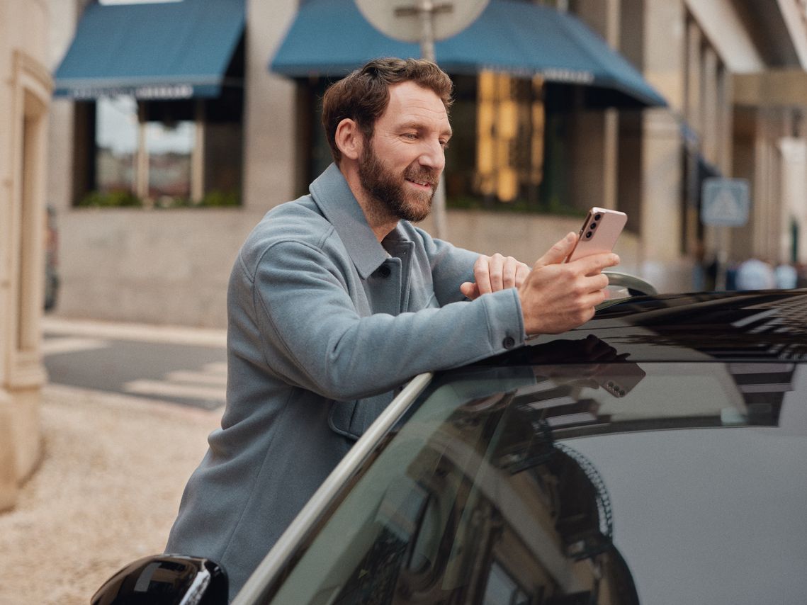 Mann lehnt an einem Volkswagen und schaut auf sein Mobiltelefon