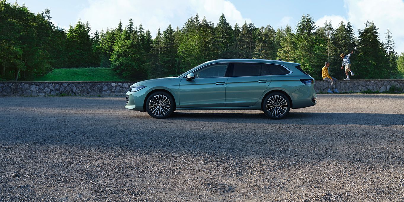 Ein grüner VW Passat B9 in der Seitenansicht, im Hintergrund zwei Menschen und Wald