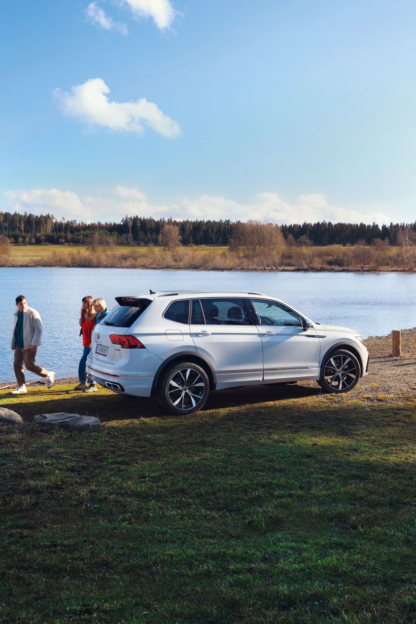 Ein weißer VW Tiguan Allspace steht geparkt auf einer Wiese vor einem See. Dahinter laufen 3 Menschen entlang.