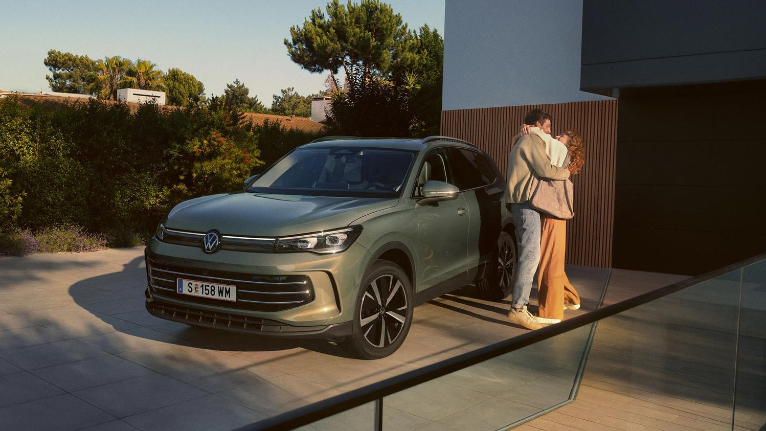 Eine Frau und ein Mann umarmen sich, neben ihnen steht ein VW Tiguan Elegance in der Farbe Cipressino-Grün Metallic