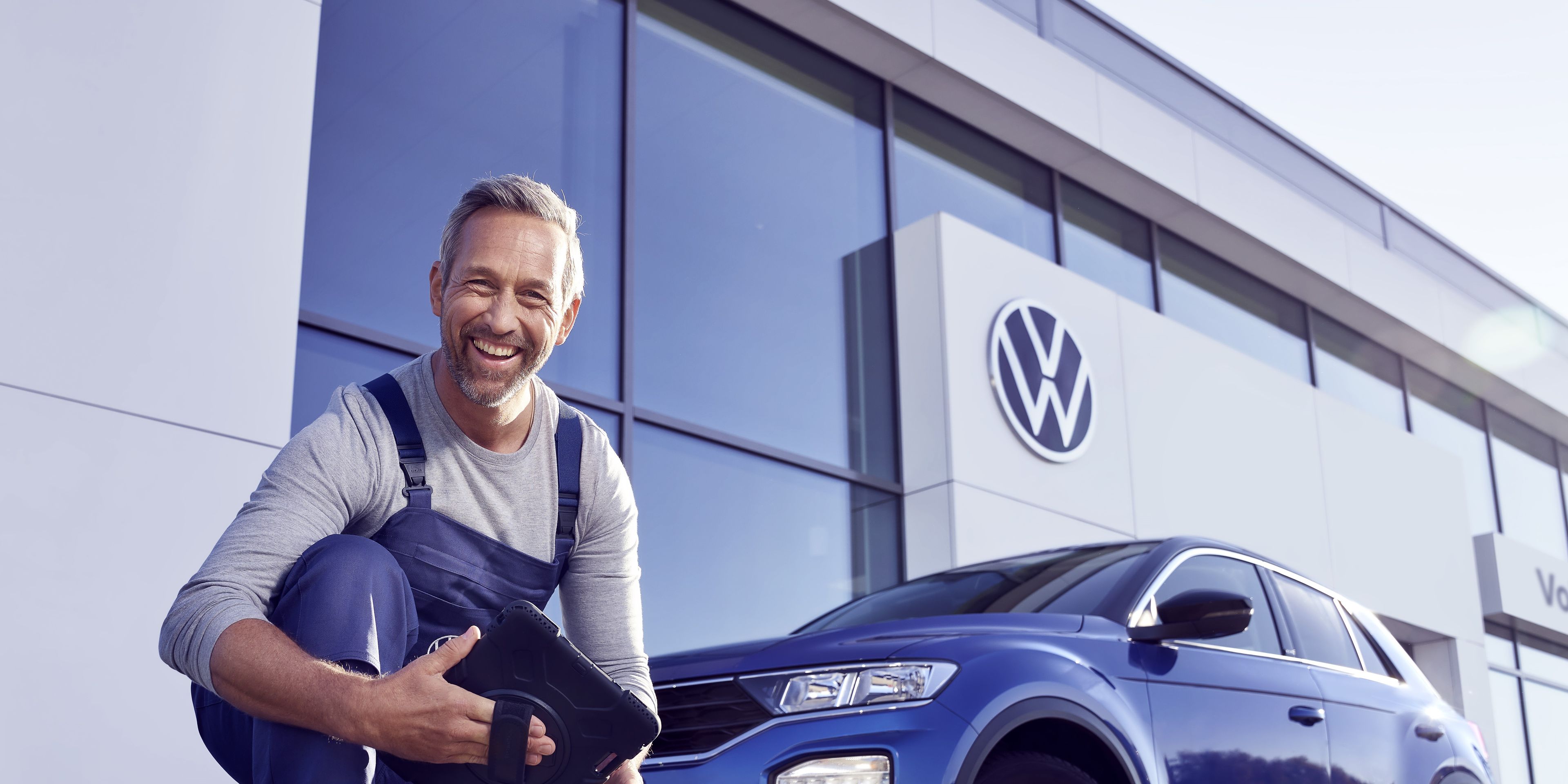 Ein Volkswagen Service Techniker mit Tablet, kniet vor einem blauen Volkswagen T-Roc