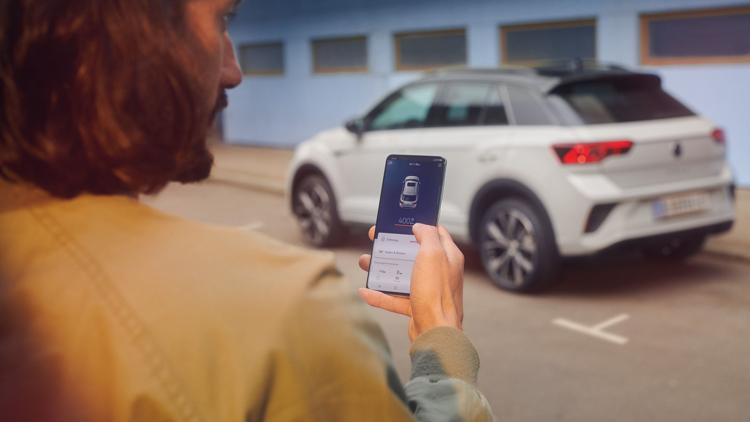 VW T-Roc R-Line in weiß vor einem Gebäude, Seite sichtbar, Mann steht mit Smartphone im Vordergrund