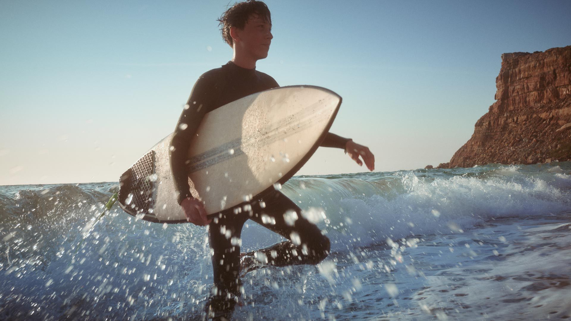 Strandszene - Ein junger Mann geht mit einen Surfbrett unter dem Arm durch das kniehohe Wasser.  