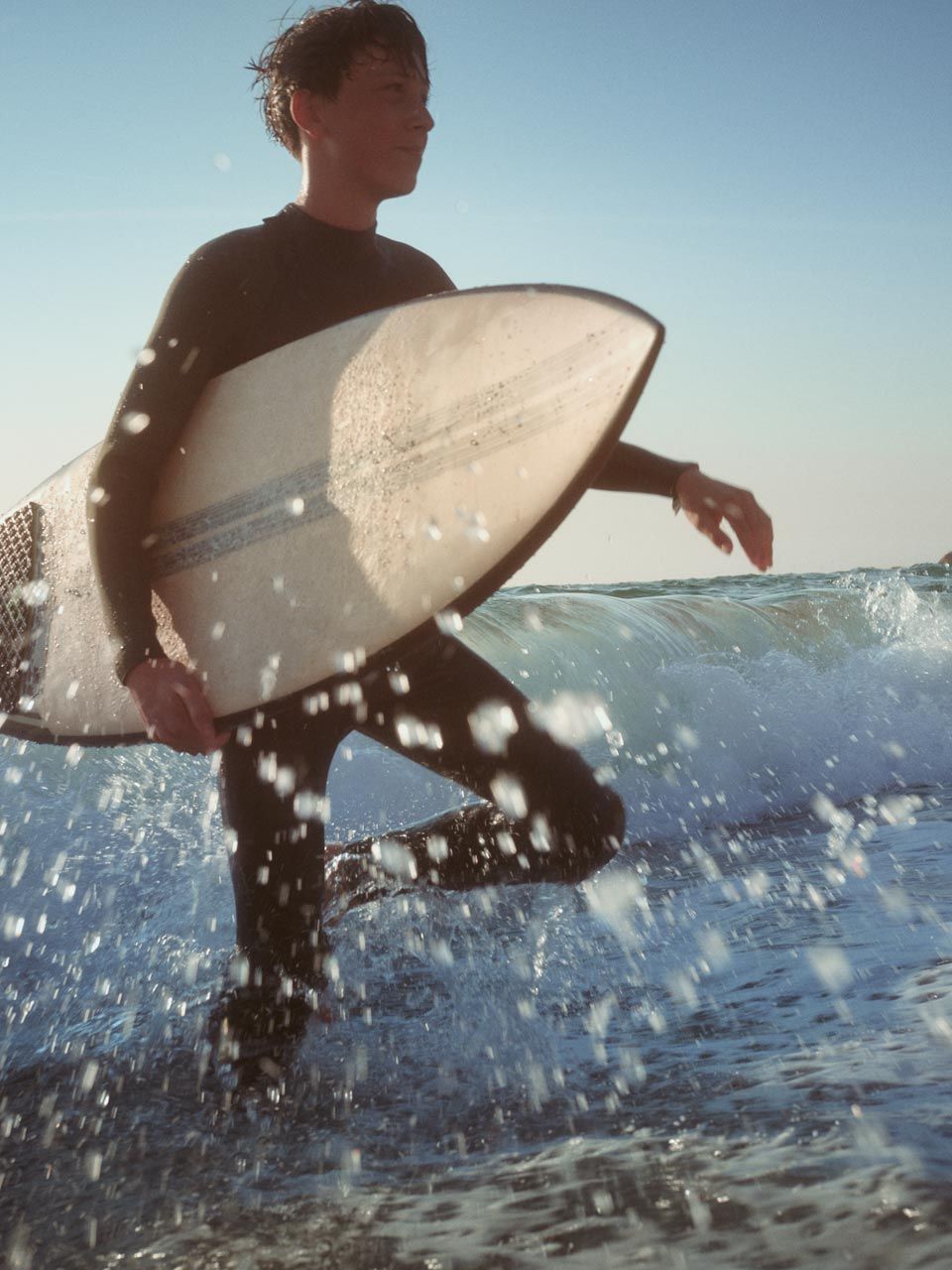 Strandszene - Ein junger Mann geht mit einen Surfbrett unter dem Arm durch das kniehohe Wasser.  