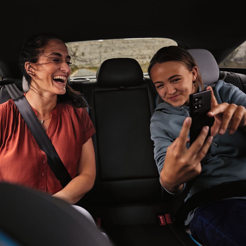 2 Peronen sitzen auf der Rückbank eines Volkswagen und schauen lächelnd auf ein Mobiltelefon