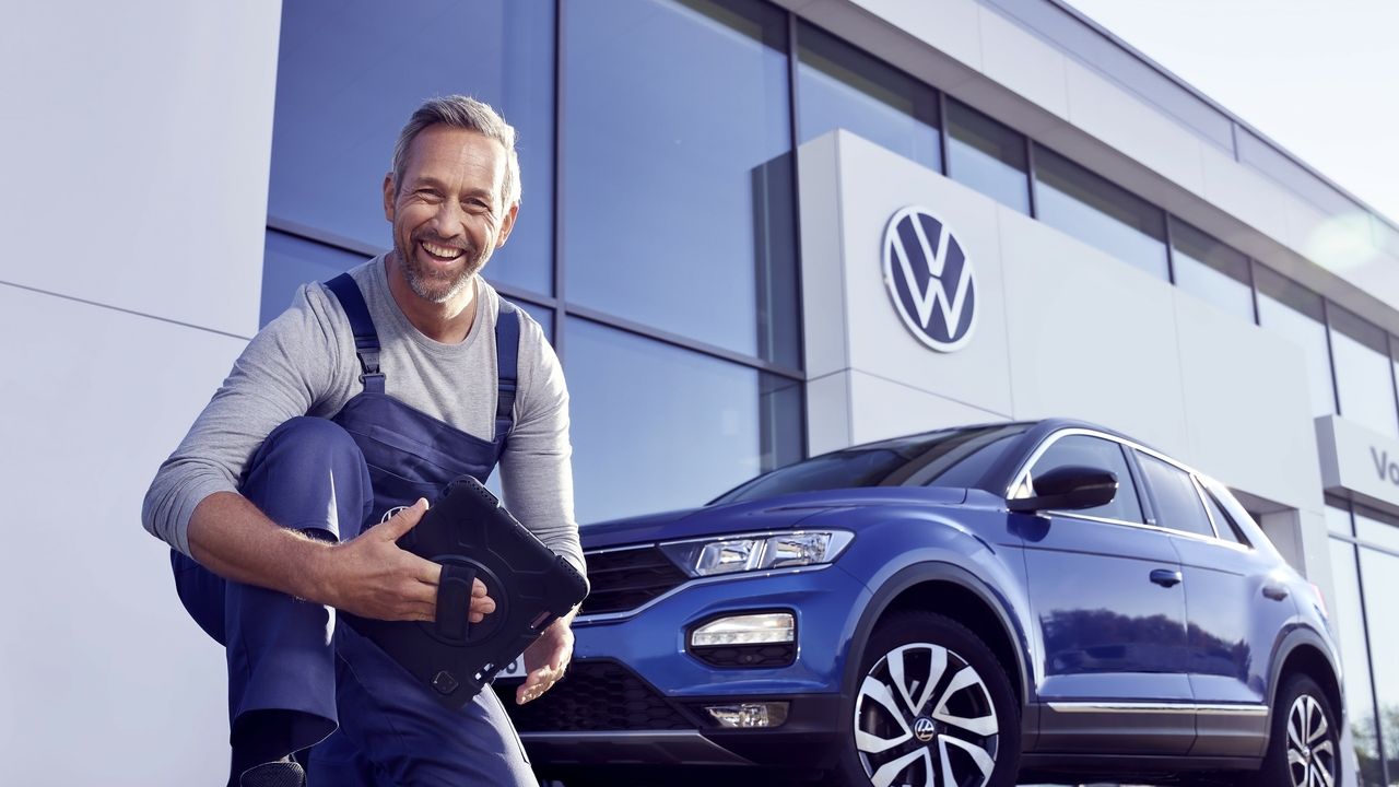 Ein Volkswagen Service Techniker mit Tablet, kniet vor einem blauen Volkswagen T-Roc