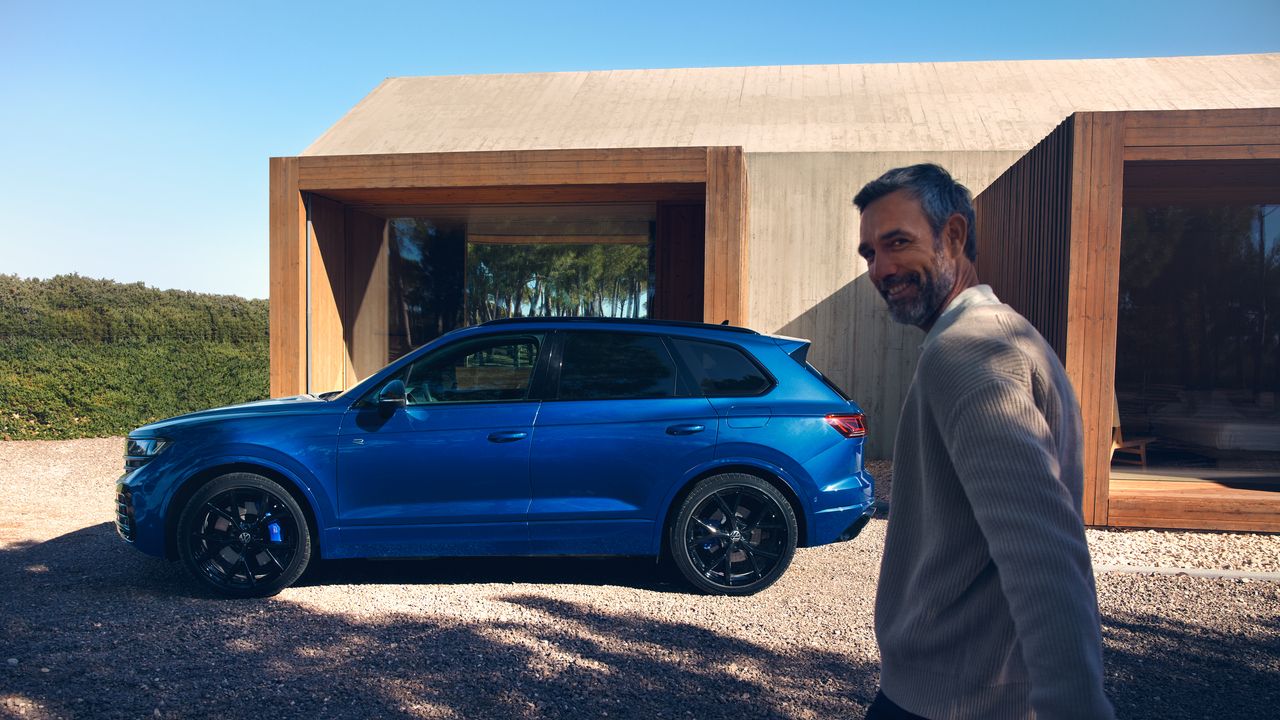 Seitenansicht des VW Touareg R eHybrid, der vor einem Haus parkt. Ein Mann geht auf das Fahrzeug zu.