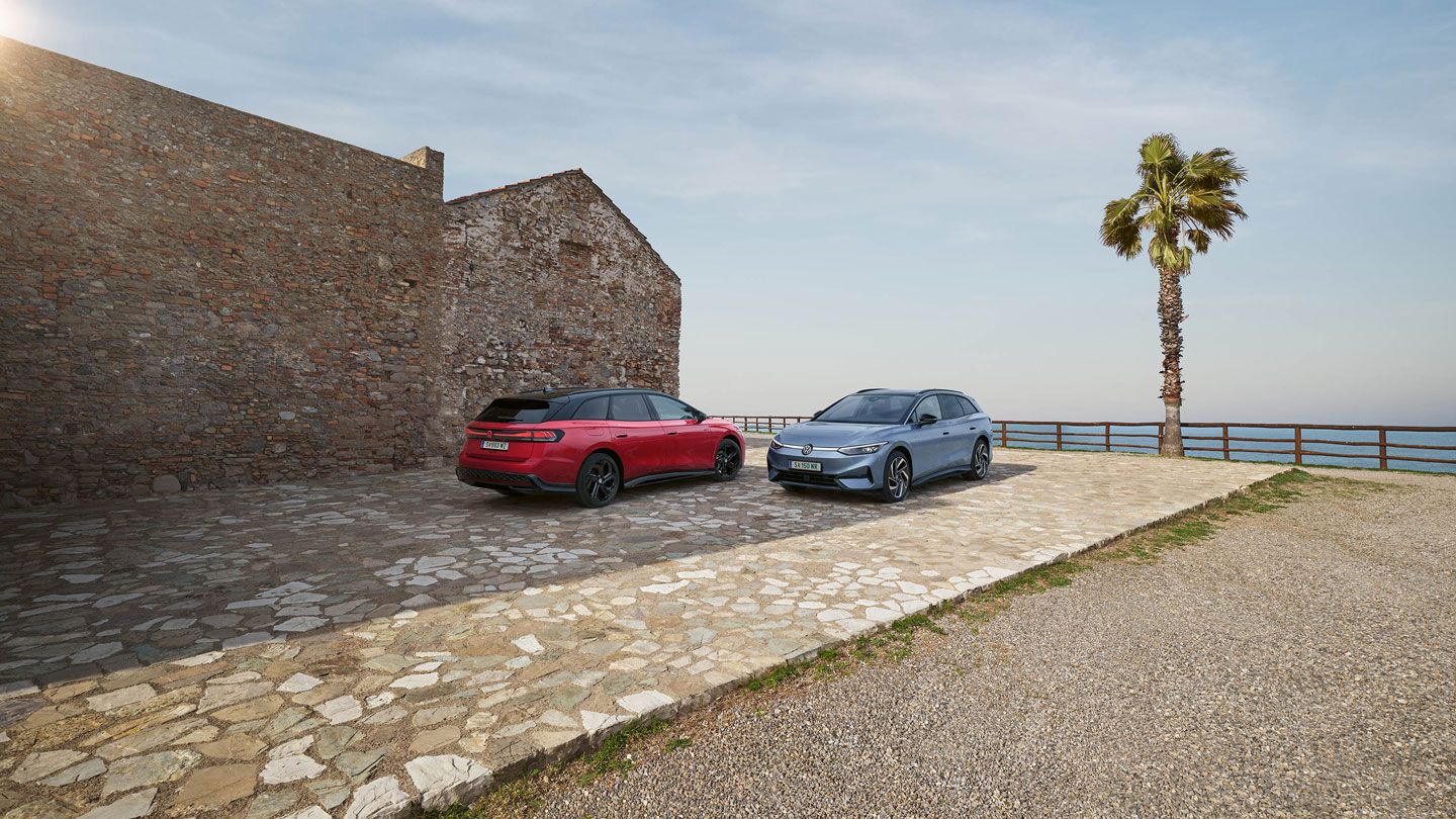 Ein VW ID.7 Tourer in Stonewashed Blue Metallic und ein VW ID.7 Tourer GTX in Kings Red Metallic geparkt an Küste neben eines Steinhaus'