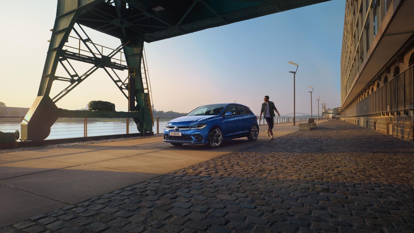  Ein blauer VW Polo R-Line steht an der Promenade neben einem Fluss, ein Mann läuft um das Auto herum.