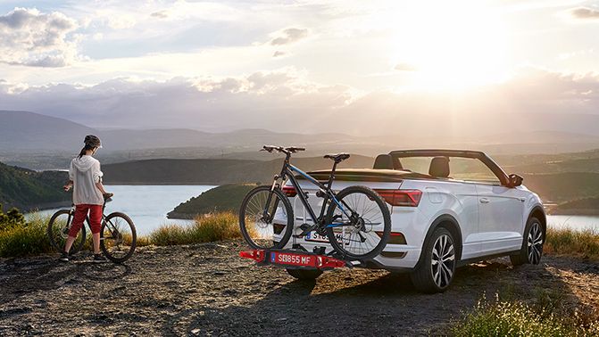 Ein VW T-Roc Cabrio in weiss mit Fahrradträger parkt in der Nähe eines Sees, eine Frau mit einem Fahrrad steht neben dem Auto.