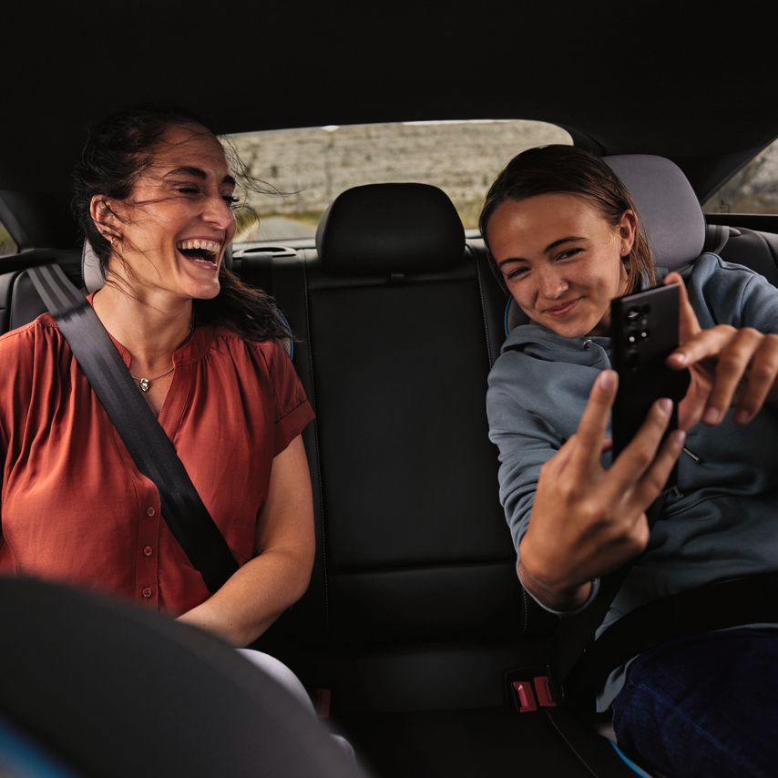 2 Peronen sitzen auf der Rückbank eines Volkswagen und schauen lächelnd auf ein Mobiltelefon