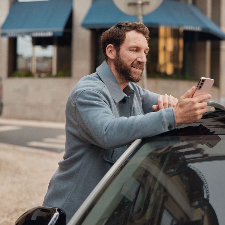 Mann lehnt an einem Volkswagen und schaut auf sein Mobiltelefon