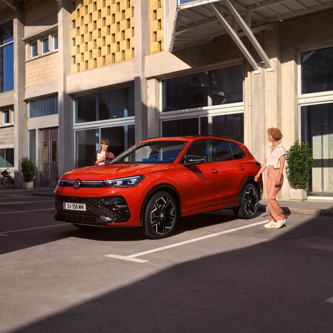 Roter VW Tiguan R-Line in Seitenansicht steht vor einem Gebäude, eine junge Frau geht auf der Fahrerseite vorbei.