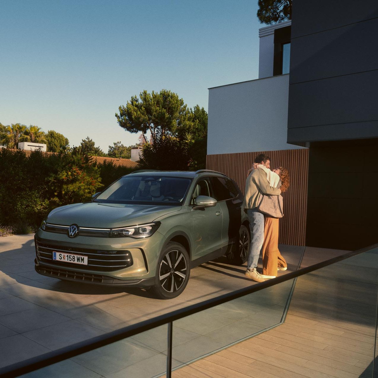 Grüner VW Tiguan parkt in Frontansicht auf der Einfahrt eines modernen Wohnhauses. Ein junges Paar steht daneben und umarmt sich.