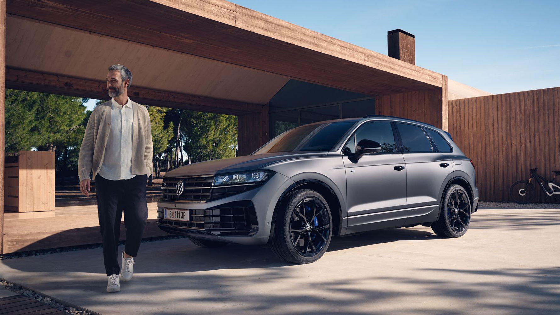 Ein VW Touareg R eHybrid parkt vor einem Haus, ein Mann bewegt sich vom Fahrzeug fort