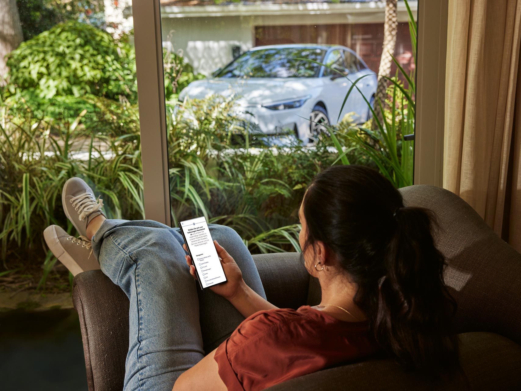 Eine Frau sitzt entspannt im Sofa und blickt auf ihr Smartphone, im Hintergrund ein VW ID.7